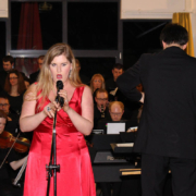 An Mikrofon in roten Abendkleid singt vor Orchester eine Sängerin links neben ihr dirigiert ein Dirigent.
