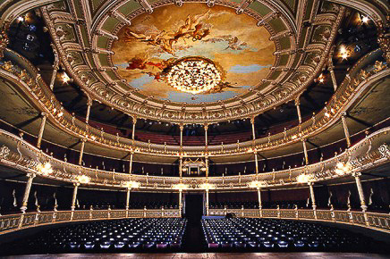 Ein grosser Opernsaal wird aus Blickwinkel der Bühne abgelichtet. Oben an der Decke befindet sich eingebettet in einer barocken Malerei ein grosser Kromleuchter. Das Theater hat 2 Ränge.