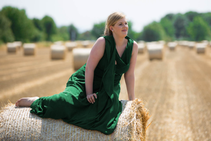 Eine blonde Frau im grünen Abendkleid blickt auf einem Heuballen in die Weite