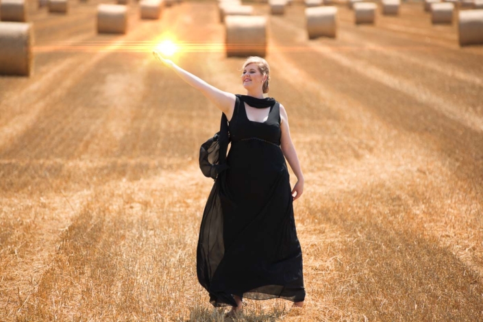 Auf einem abgeernten Getreidefeld steht eine Frau im schwarzen Abendkleid und hält auf ausgestreckter rechter Hand einen virtuellen Feuerball