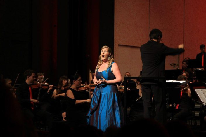 Eine Opernsängerin singt im hellblauen Abendkleid links vom Dirigenten vor grossem Orchester in einem Konzertsaal