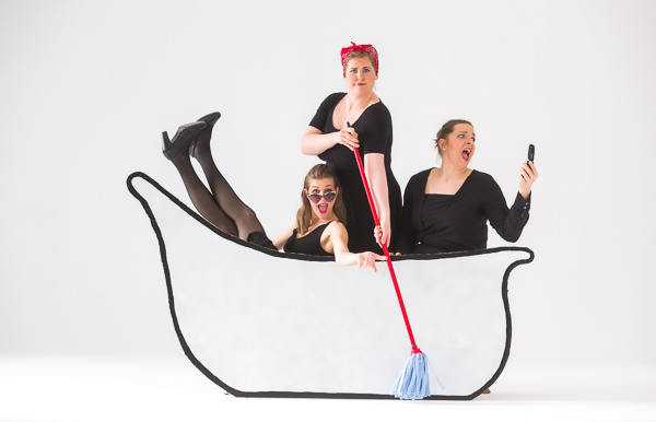 Drei Frauen in schwarzer Kleidung sitzen in einer Pappbadewanne und schauen differenziert theatralisch in die Kamera