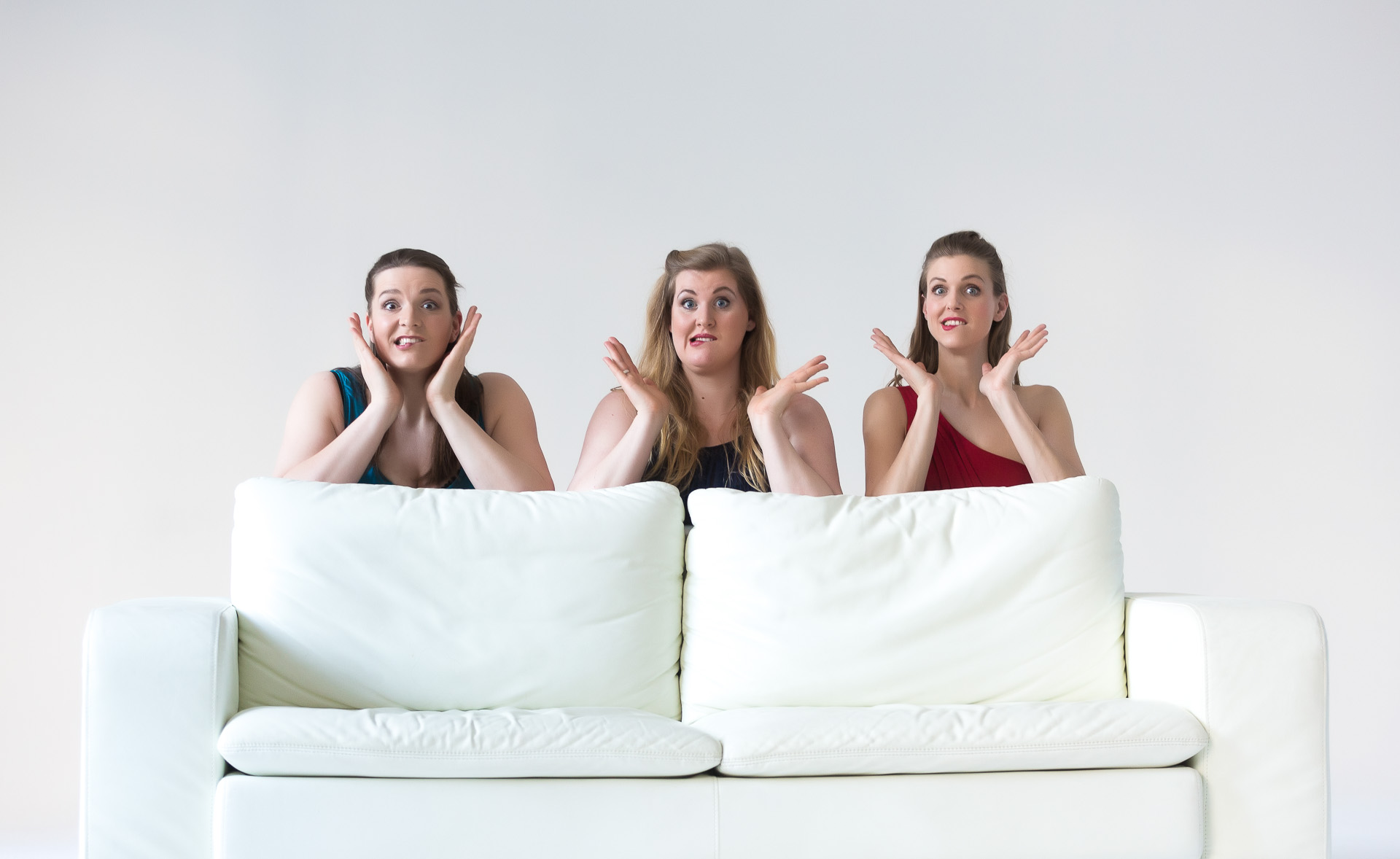 Drei Frauen im Abendkleid ziehen knieend hinter einer weissen Couch Grimassen